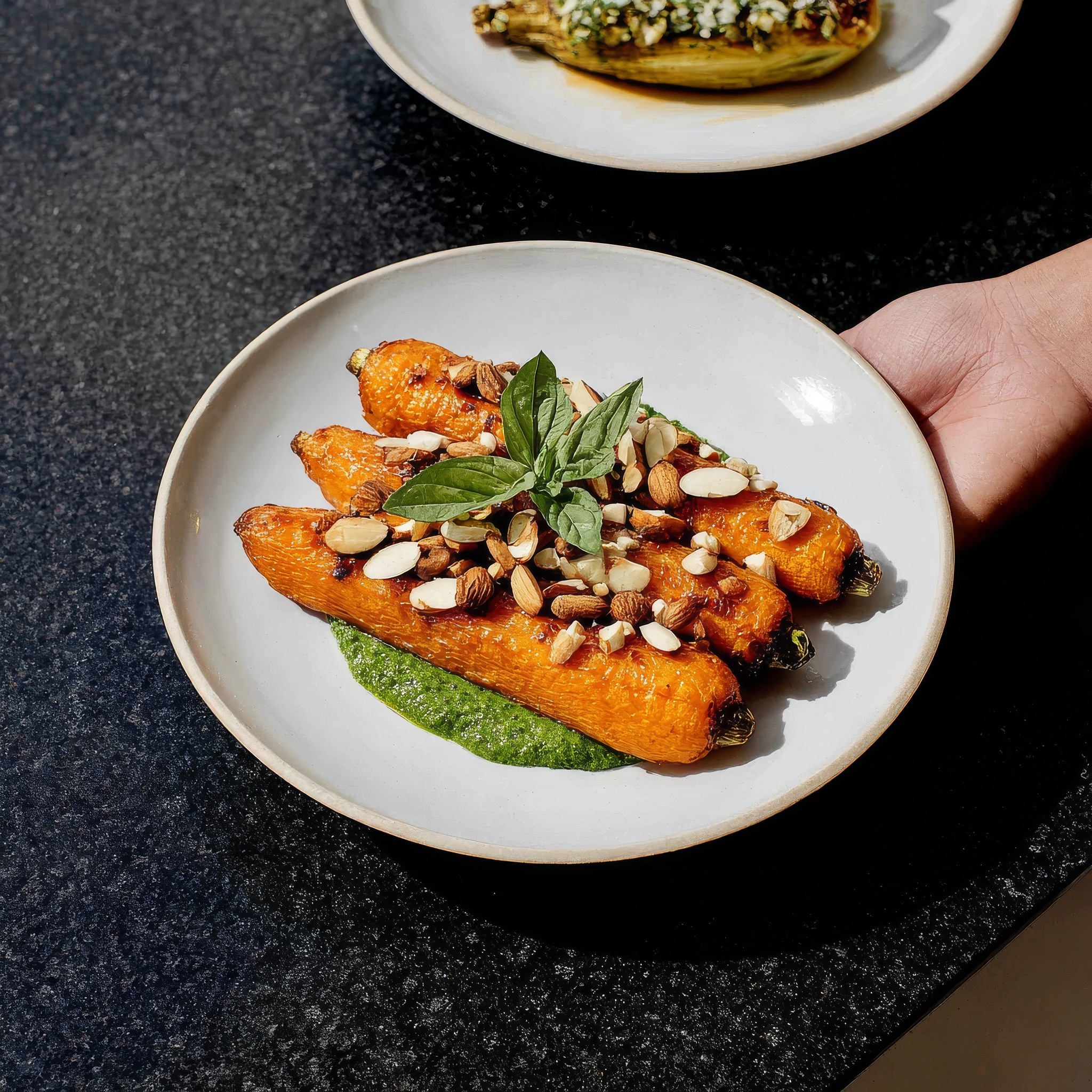 White plate with roasted carrots served on green sauce, topped with almonds and fresh herbs.