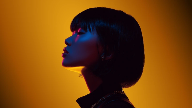 Profile of a woman with short hair illuminated by blue and pink lighting against an orange background.